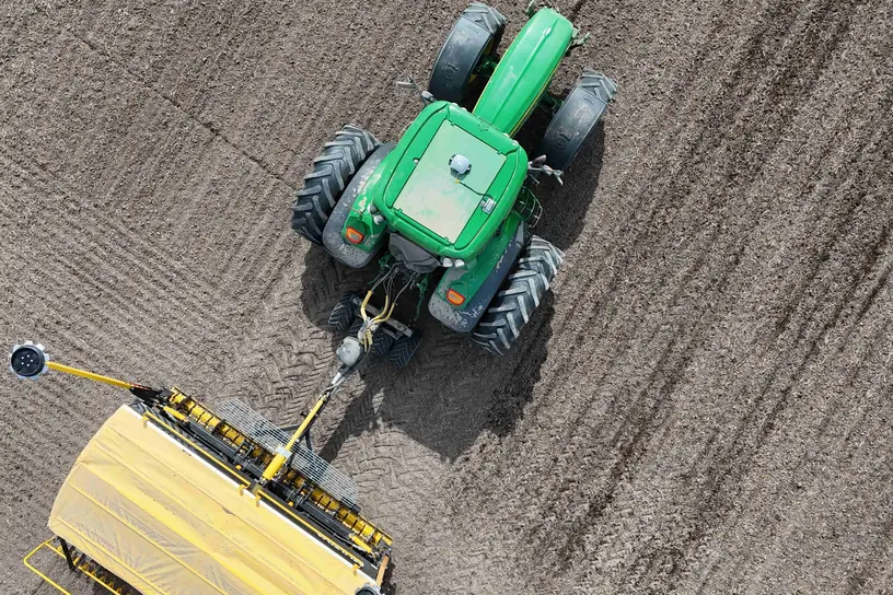 Vista zenital d'un tractor verd estirant un aper groc sobre un camp llaurat