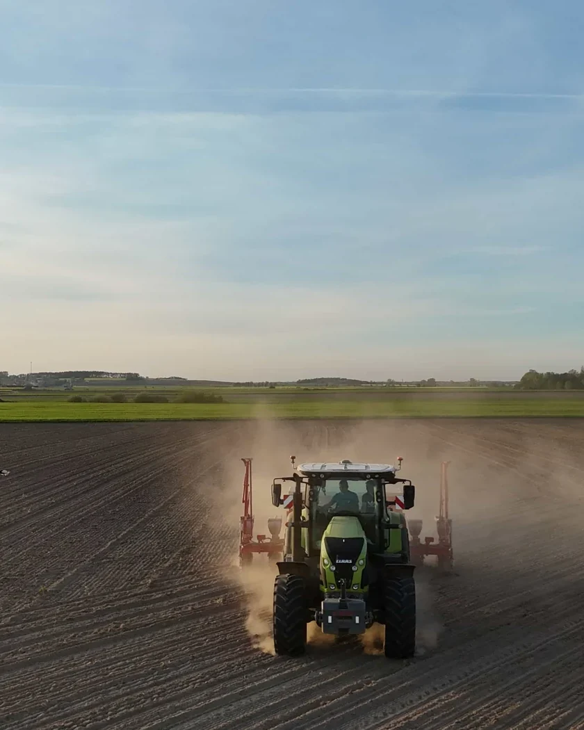 Tractor sembrant un camp llaurat pla, vist de cara des de l'aire, amb una estela de pols i el cel clar del capvespre