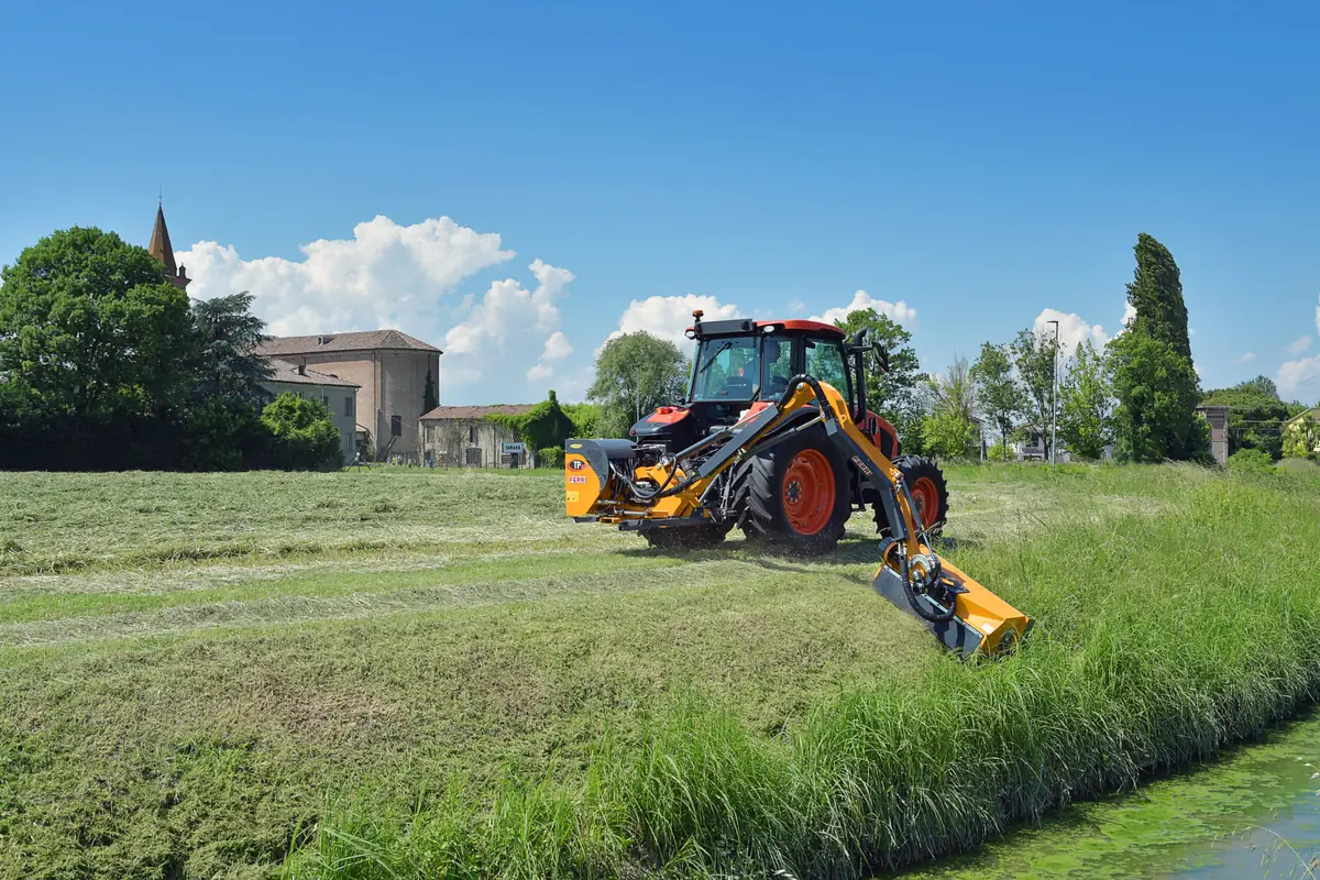 Tractor taronja amb un braç desbrossador Ferri segant el marge herbós d'un canal, amb un edifici de pedra i un campanar al fons