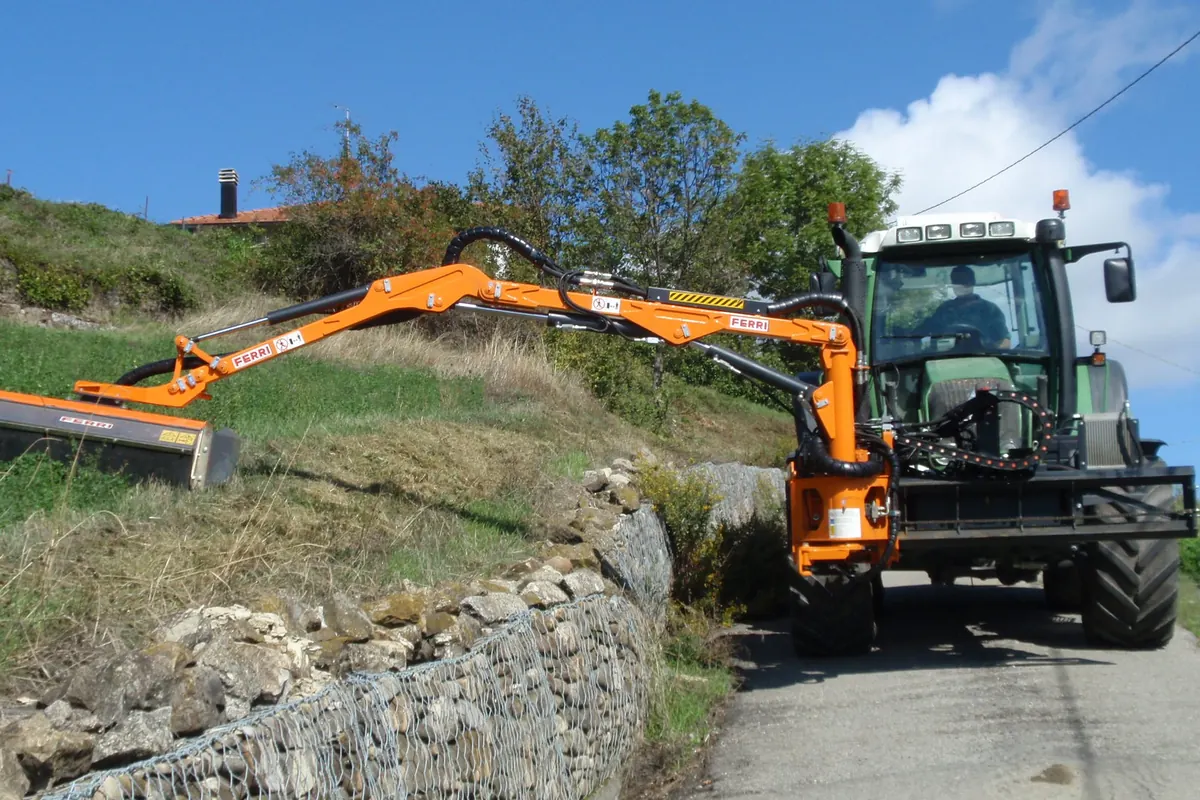 Braç desbrossador taronja muntat al davant d'un tractor verd, segant el talús d'una carretera estreta, damunt d'un mur de gabions de pedra