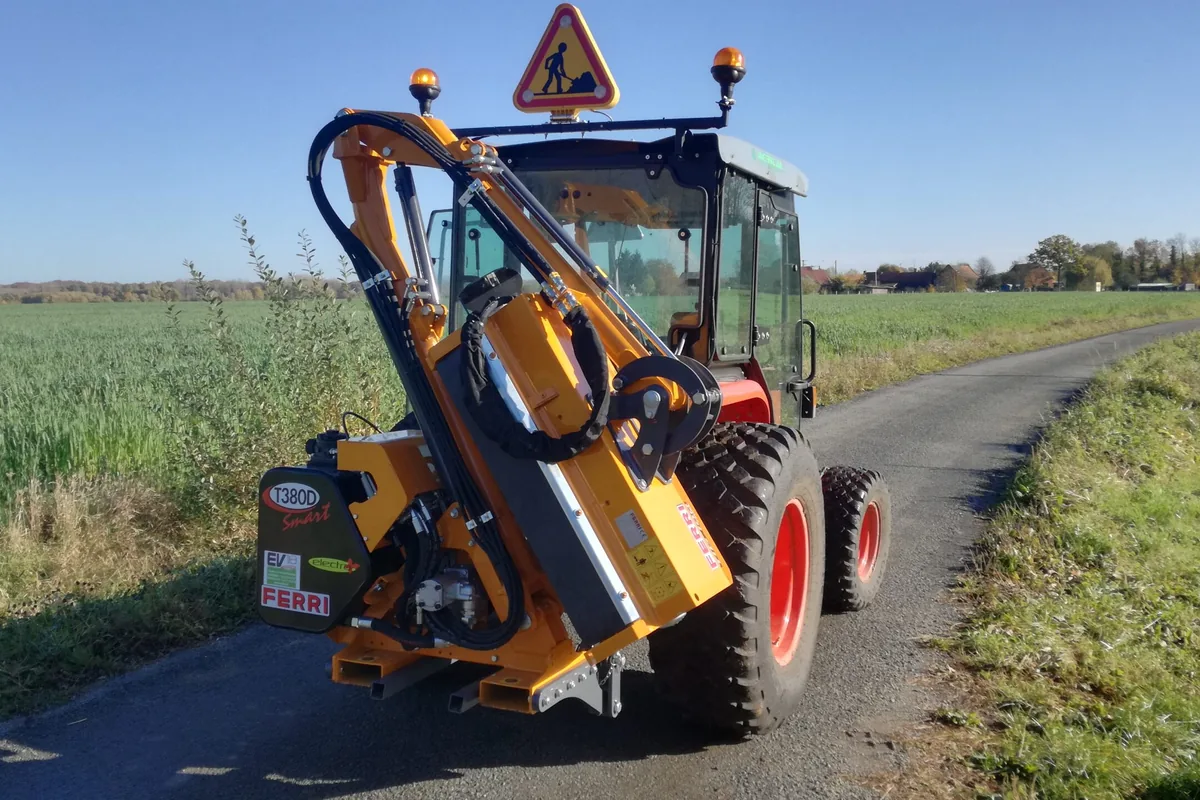Braç desbrossador Ferri T 380 D plegat en posició de transport al darrere d'un tractor de rodes vermelles, en una carretera estreta entre camps