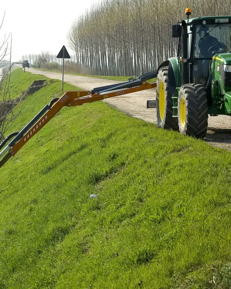 Tractor verd amb un braç desbrossador Ferri estirat cap avall, segant l'herba d'un marge amb arbres, sota el cel