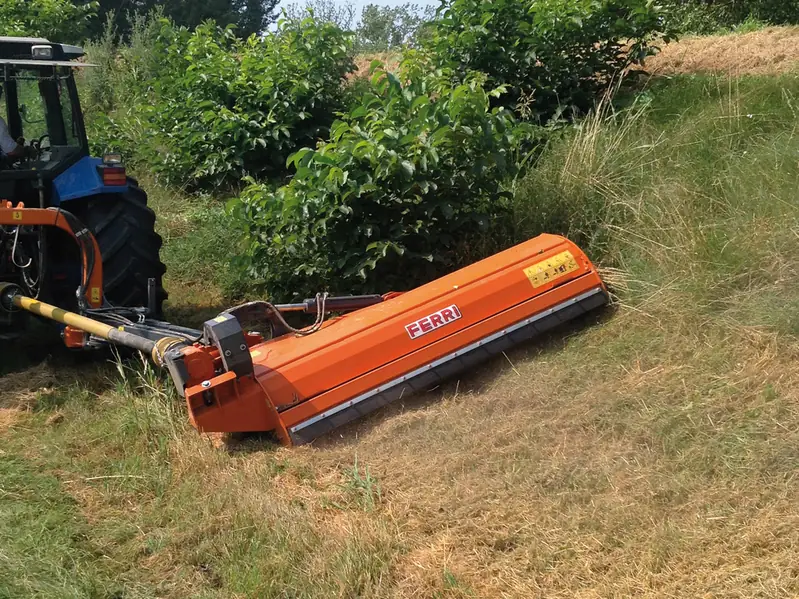 Desbrossadora lateral Ferri taronja segant un marge amb matolls darrere d'un tractor blau
