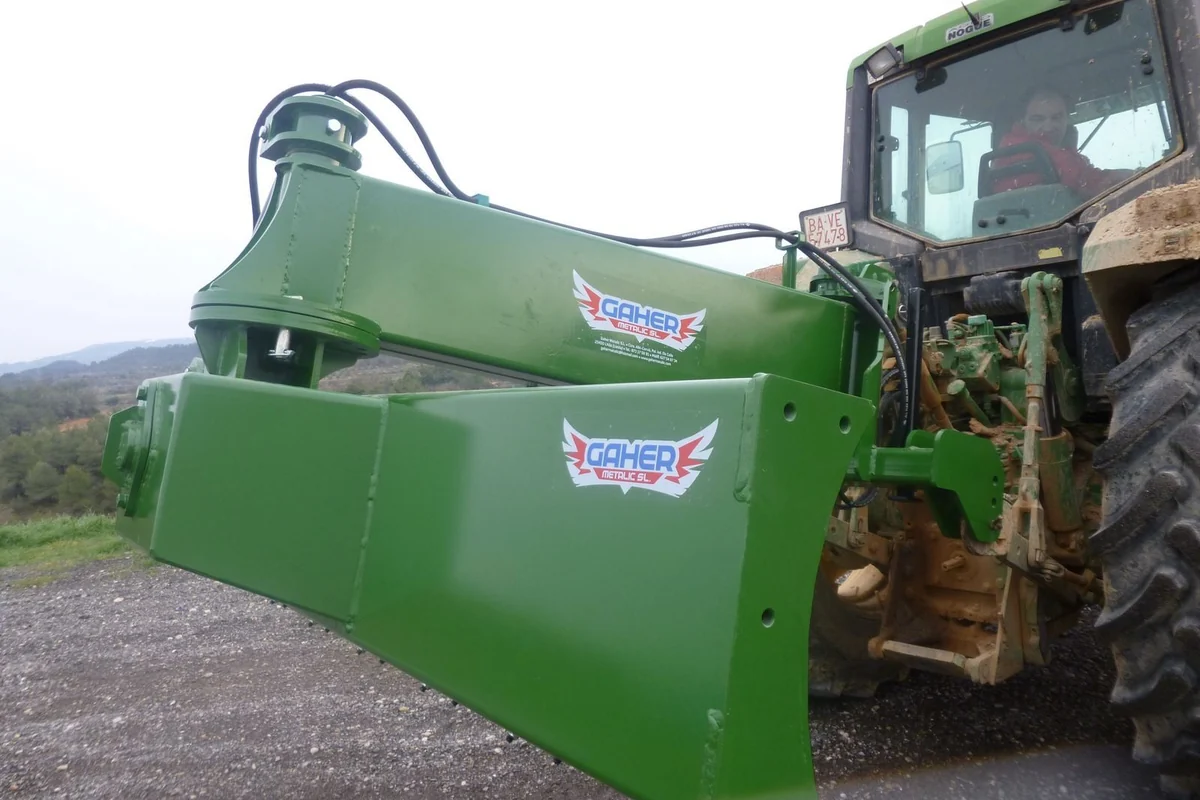 Braç i pala d'un polidozer posterior Gaher vistos pel darrere, muntats als tres punts d'un tractor verd sobre un camí de grava, amb turons al fons