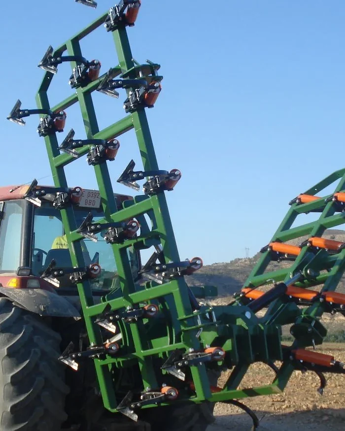 Semixissel Gaher amb una ala aixecada en vertical darrere d'un tractor vermell, en un camp llaurat, amb turons i el cel blau al fons