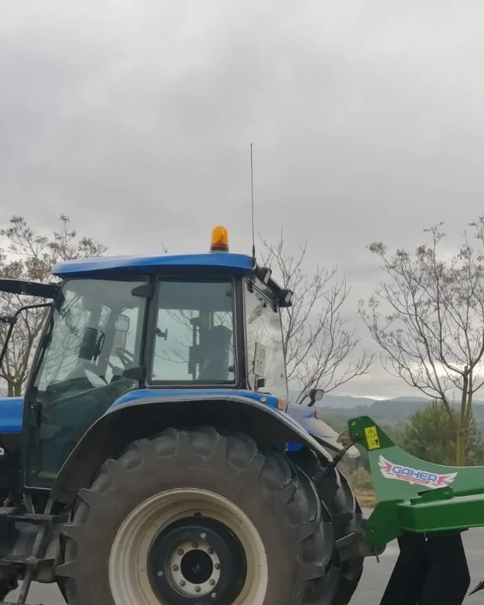 Tractor blau amb un subsolador Gaher enganxat al darrere, de perfil sobre asfalt, amb arbres sense fulles i el cel ennuvolat