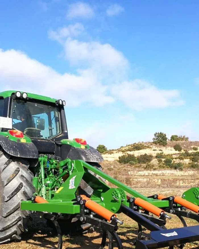 Tractor verd amb un xissel Gaher enganxat al darrere en un camp sec, amb el cel blau i núvols blancs al damunt