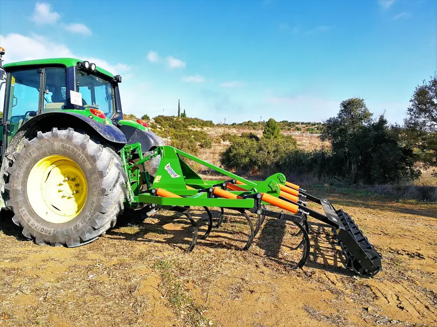 Tractor verd amb un xissel Gaher enganxat al darrere, en un camp sec amb matolls