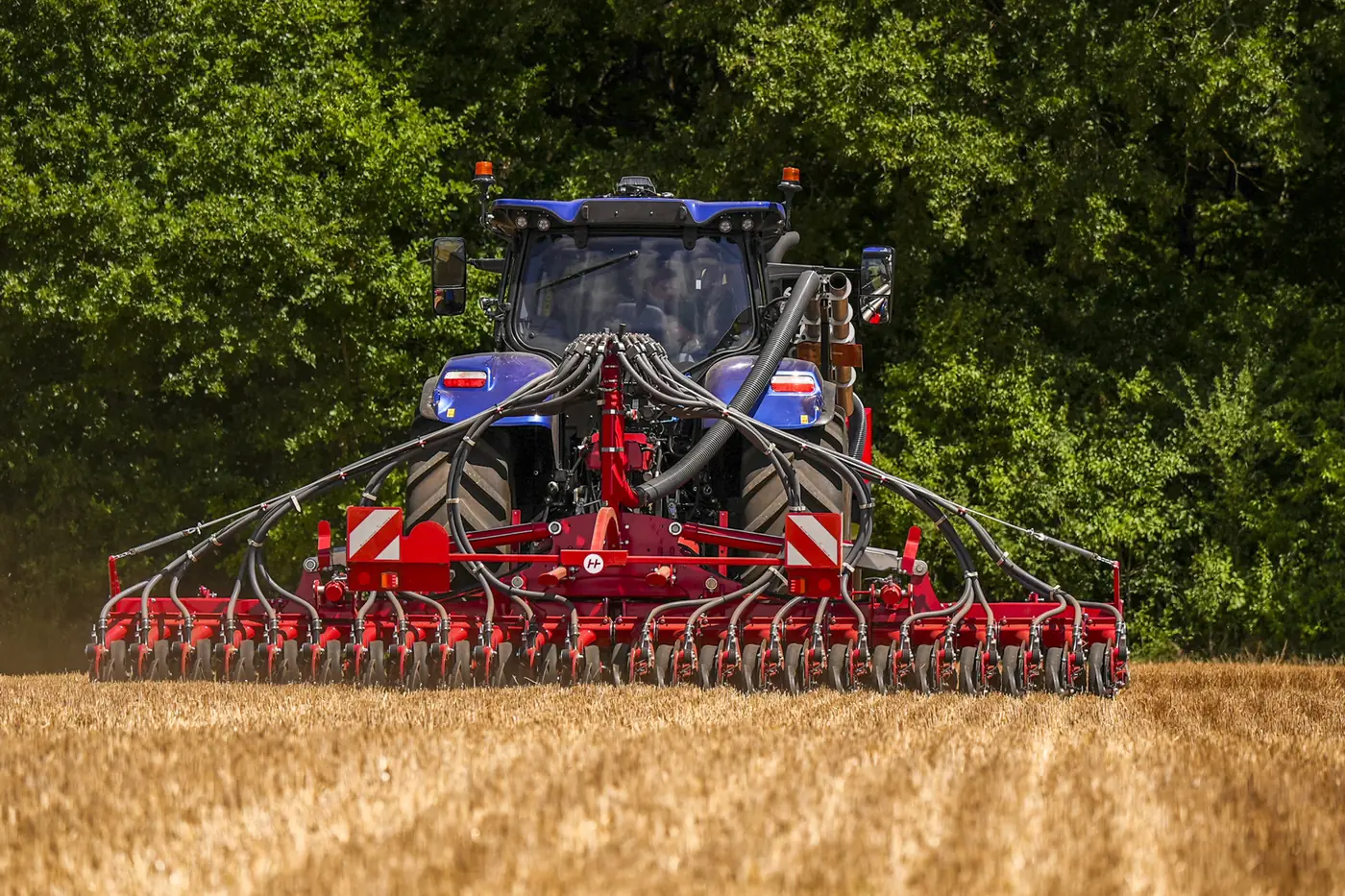 Vista des del darrere d'un tractor blau amb la barra de relles de l'Avatar enganxada al darrere, sobre rostoll i amb el bosc al fons