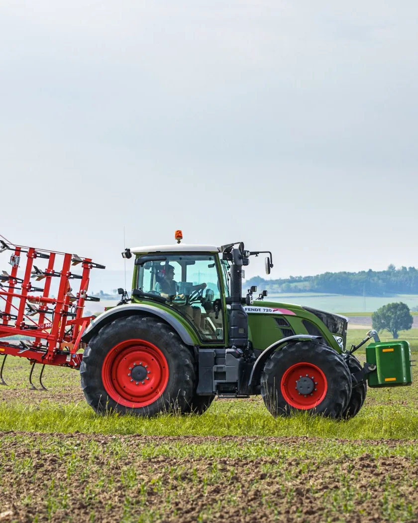 Cultivador Horsch Cruiser plegat i aixecat als tres punts d'un tractor verd, travessant un camp de cultiu acabat de néixer, amb els turons al fons sota un cel ennuvolat