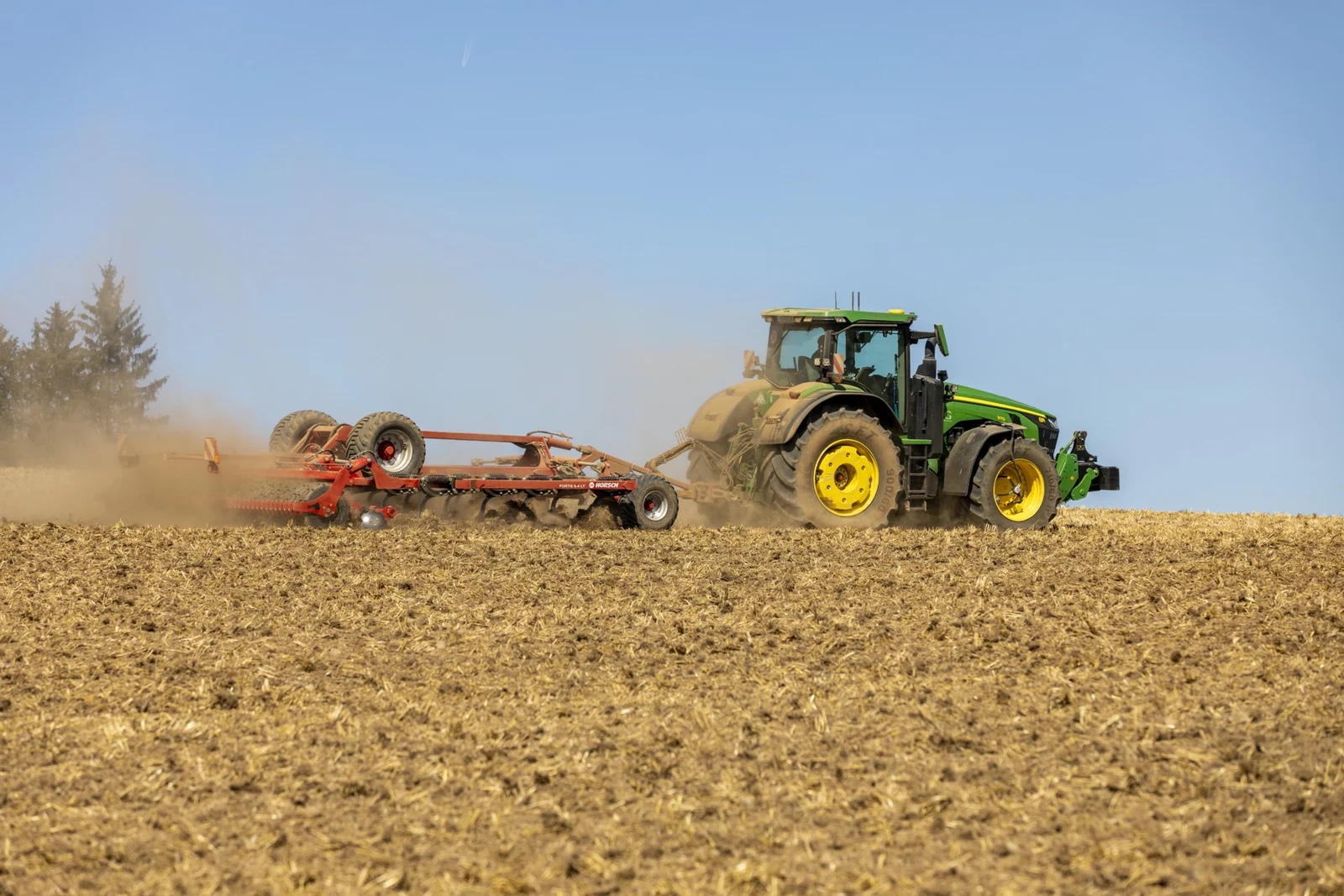 Cultivador Horsch Fortis aixecant pols darrere d'un tractor verd, en un camp acabat de treballar