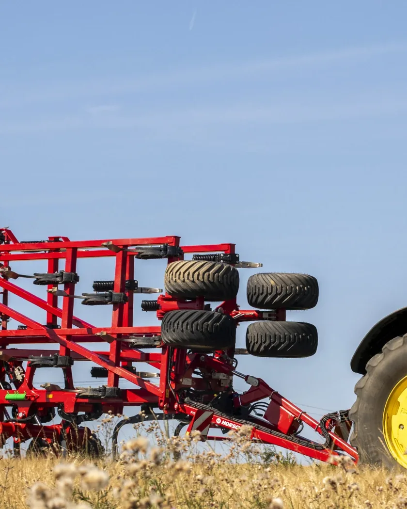Cultivador Horsch Fortis plegat i enganxat a un tractor verd, vist de costat, amb herba seca i cards en primer terme sota un cel blau