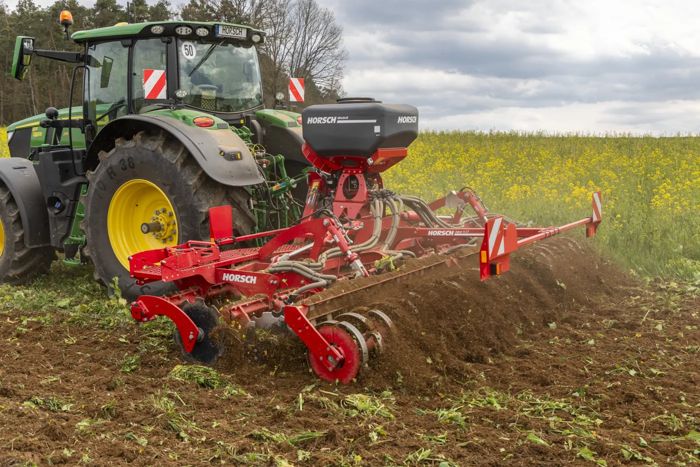 Grada de discos Horsch Joker 5 CT amb la tremuja MiniDrill al damunt, treballant un camp verd al costat d'un cultiu de flors grogues, amb el nom del model llegible