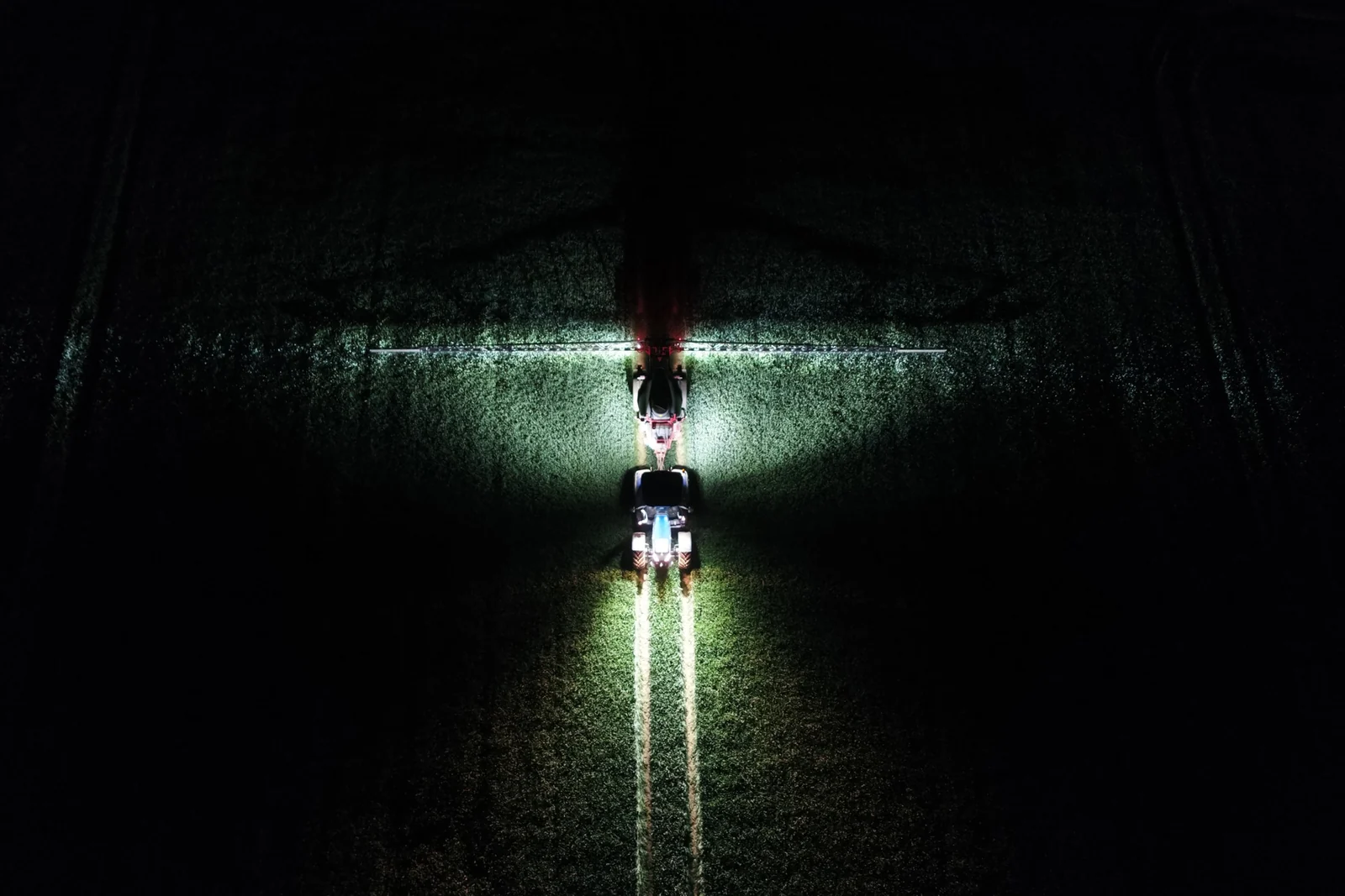 Vista aèria de nit d'un tractor amb un polvoritzador arrossegat, amb la barra desplegada i els llums il·luminant el cultiu a banda i banda