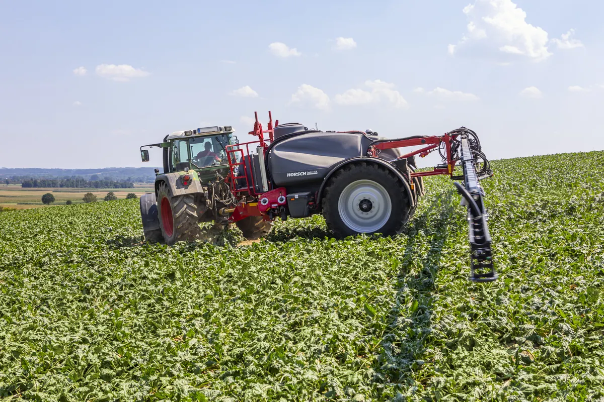 Polvoritzador arrossegat Horsch Leeb 4 AX darrere d'un tractor verd en un camp de fulla ampla, amb la barra plegada al darrere