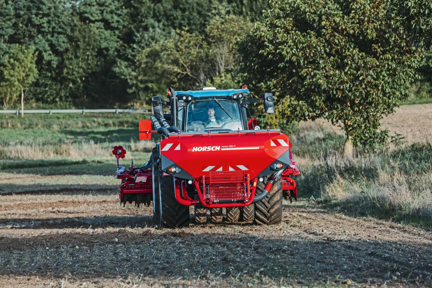 Tractor blau amb la tremuja Partner al davant, treballant al costat d'una filera d'arbres