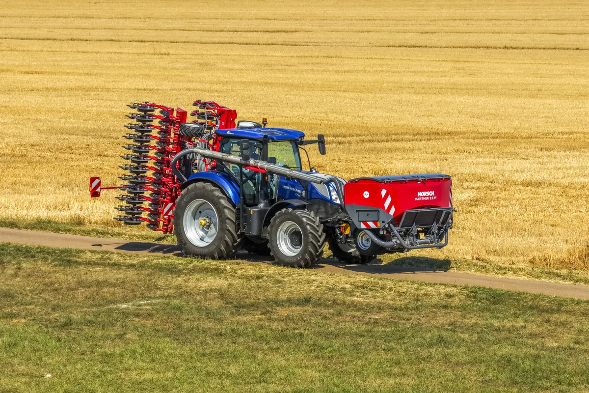 Tractor blau amb la tremuja Partner al davant i una màquina de discos plegada al darrere, vist de costat en un camí al costat d'un camp segat