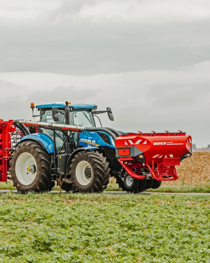 Tractor blau amb una tremuja davantera Horsch Partner i una màquina plegada al darrere, circulant per una carretera entre camps
