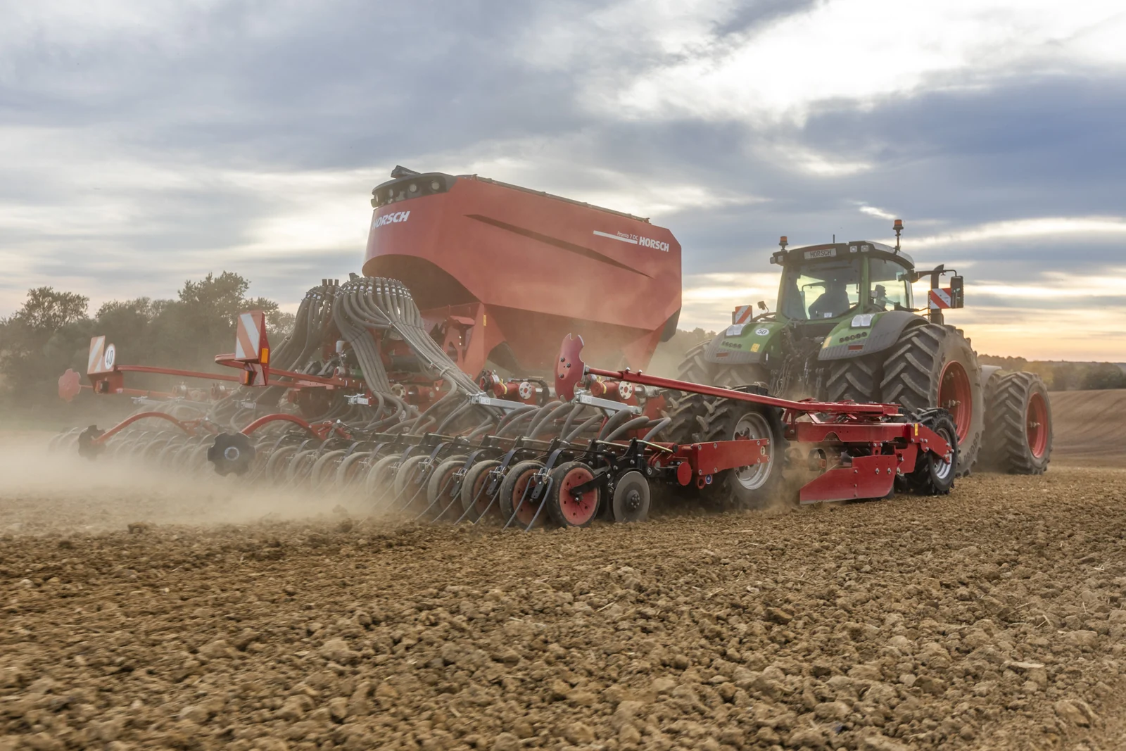 Sembradora Horsch Pronto DC vista de tres quarts, sembrant entre la pols amb la tremuja gran i un tractor verd al capdavant, sota un cel ennuvolat