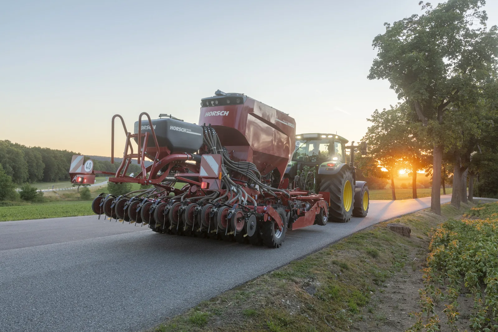 Sembradora Horsch Pronto vista des del darrere, circulant per una carretera al capvespre darrere d'un tractor verd amb els llums encesos