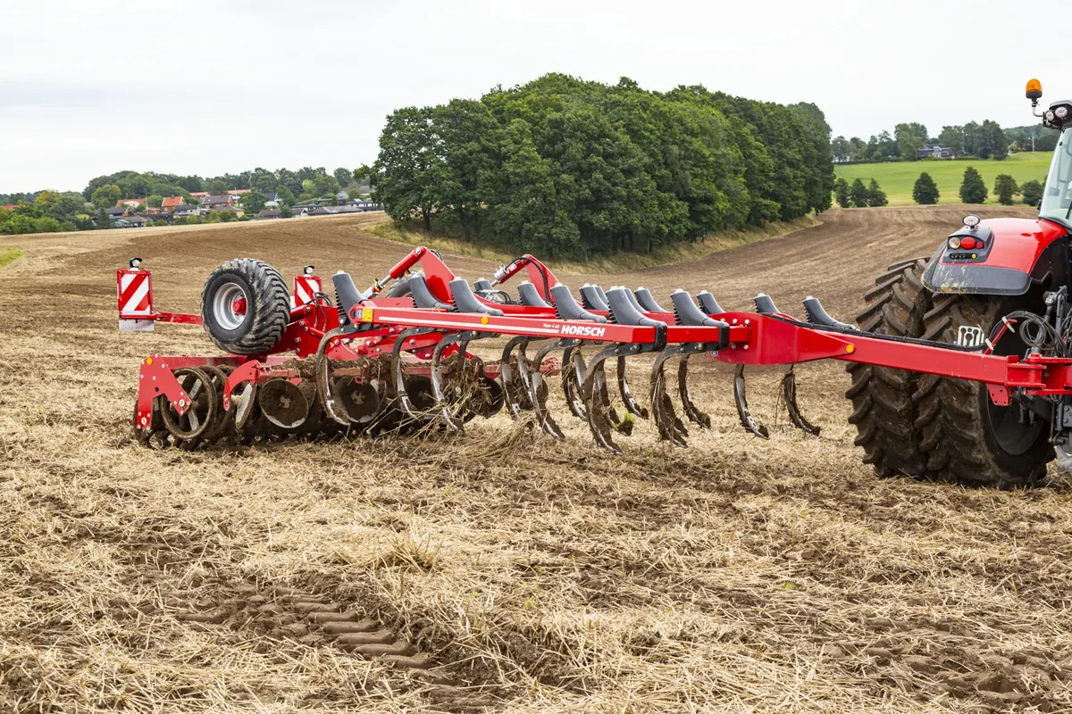 Cultivador Horsch Tiger 4 AS vist de costat sobre rostoll, amb les pues, els discos i el corró a la vista i un bosquet al fons