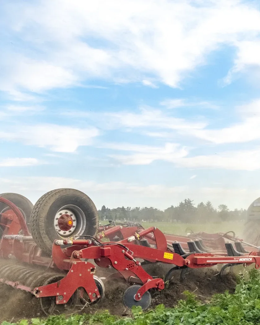Cultivador Horsch Tiger treballant un camp amb pols darrere d'un tractor, sota un cel blau amb núvols