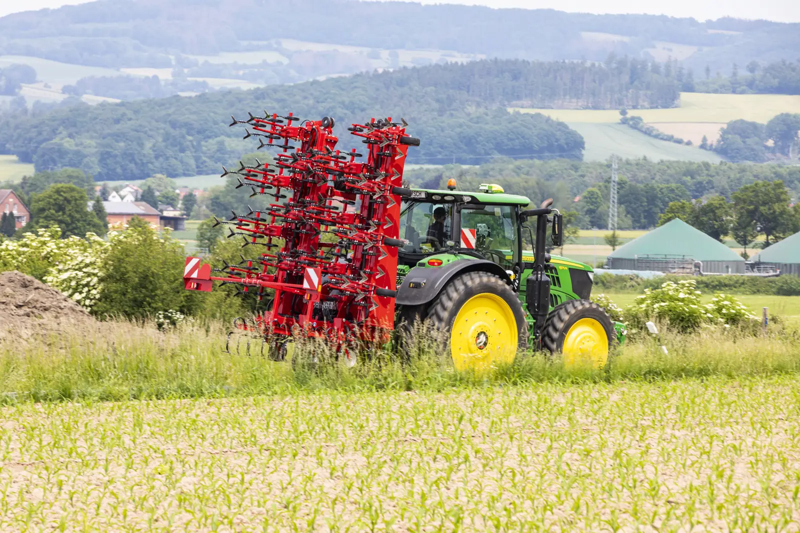 Tractor verd amb la Transformer plegada en vertical, passant per un marge d'herba al costat d'un camp de blat de moro, amb turons i boscos al fons
