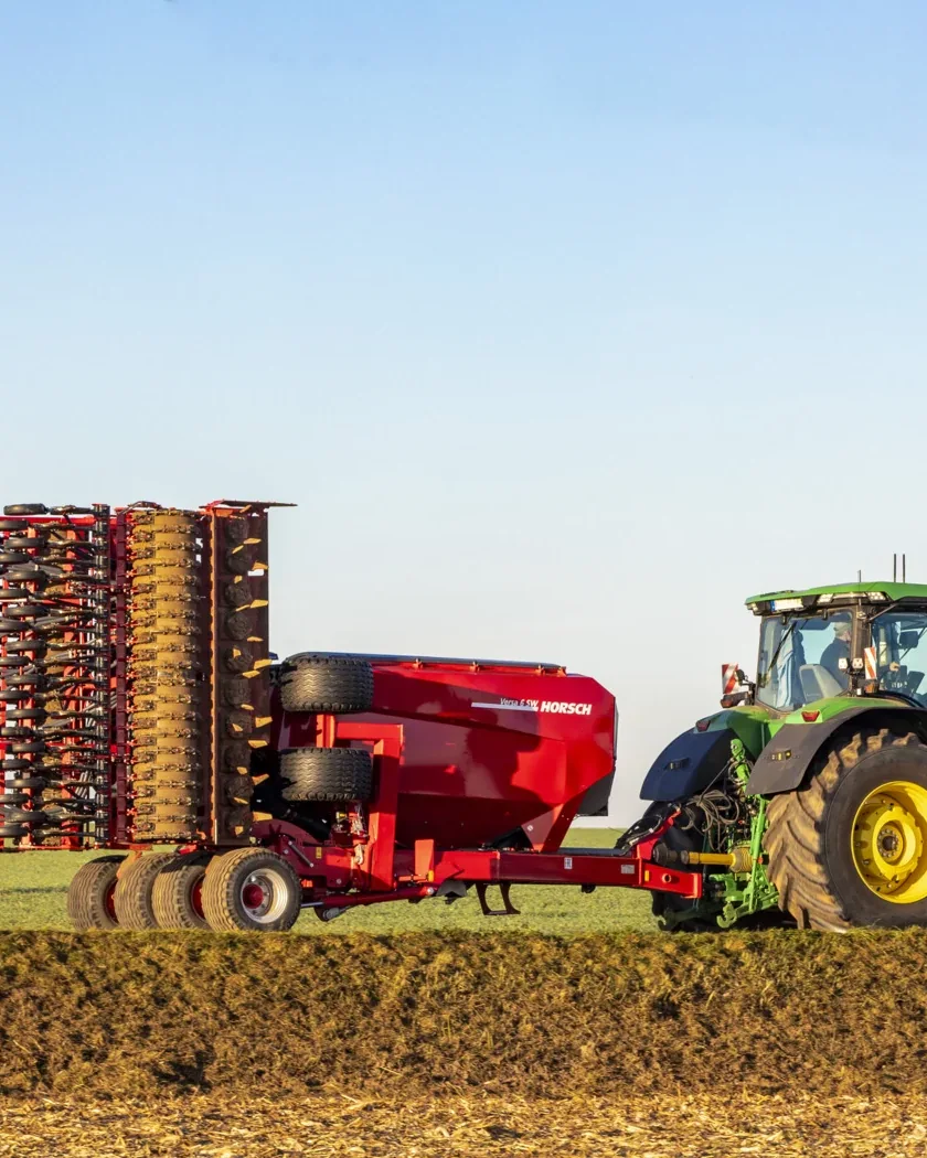 Grada rotativa Horsch Versa plegada per al transport, amb la tremuja arrossegada vermella, darrere d'un tractor verd de rodes grogues a la vora d'un camp treballat