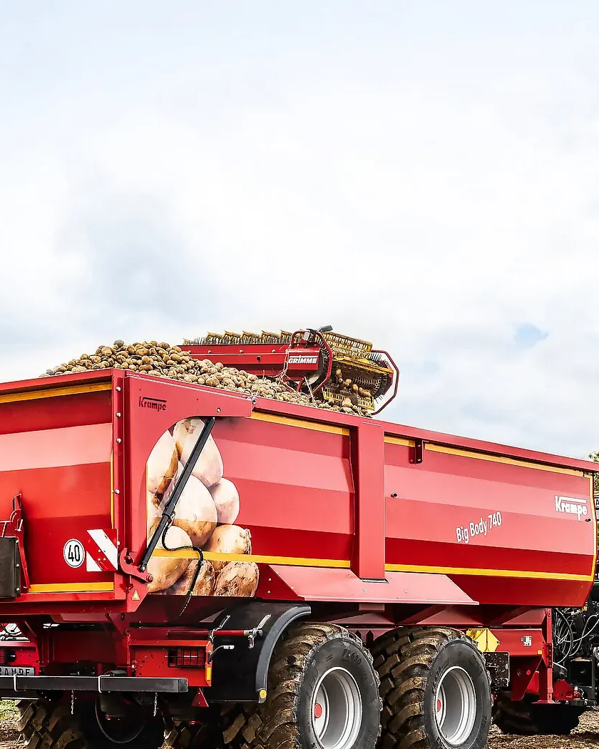 Banyera Krampe BigBody 740 carregada de patates, de tres quarts pel darrere, enganxada a un tractor en un camí amb arbres