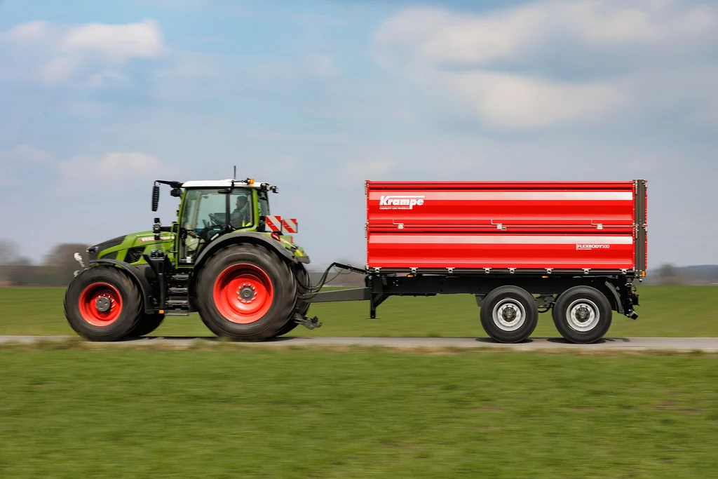 Tractor amb les llantes vermelles estirant un remolc basculador trilateral Krampe de dos eixos en tàndem per un camí, de perfil