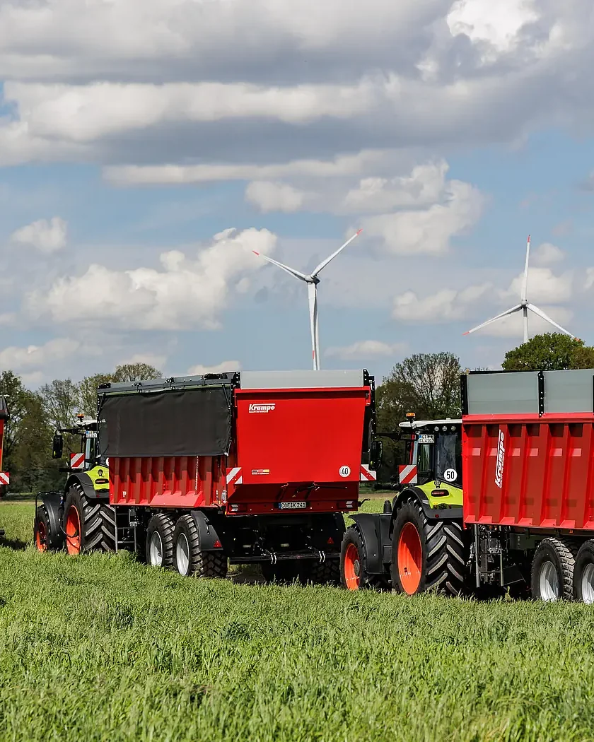 Tres remolcs Krampe RamBody en filera per un prat verd, cadascun darrere d'un tractor, amb aerogeneradors i núvols al fons