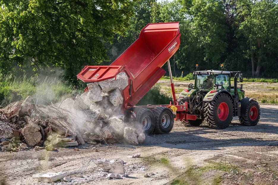 Bolquet Krampe amb la caixa aixecada abocant runa de formigó enmig d'un núvol de pols, amb el tractor a la dreta