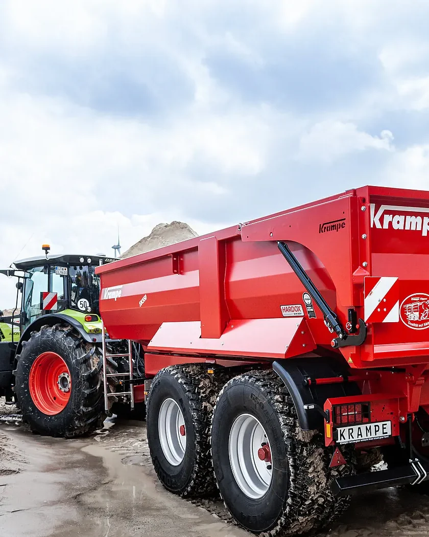 Tractor verd clar amb un bolquet Krampe SK 550 de dos eixos sobre sorra mullada, amb un aerogenerador al fons