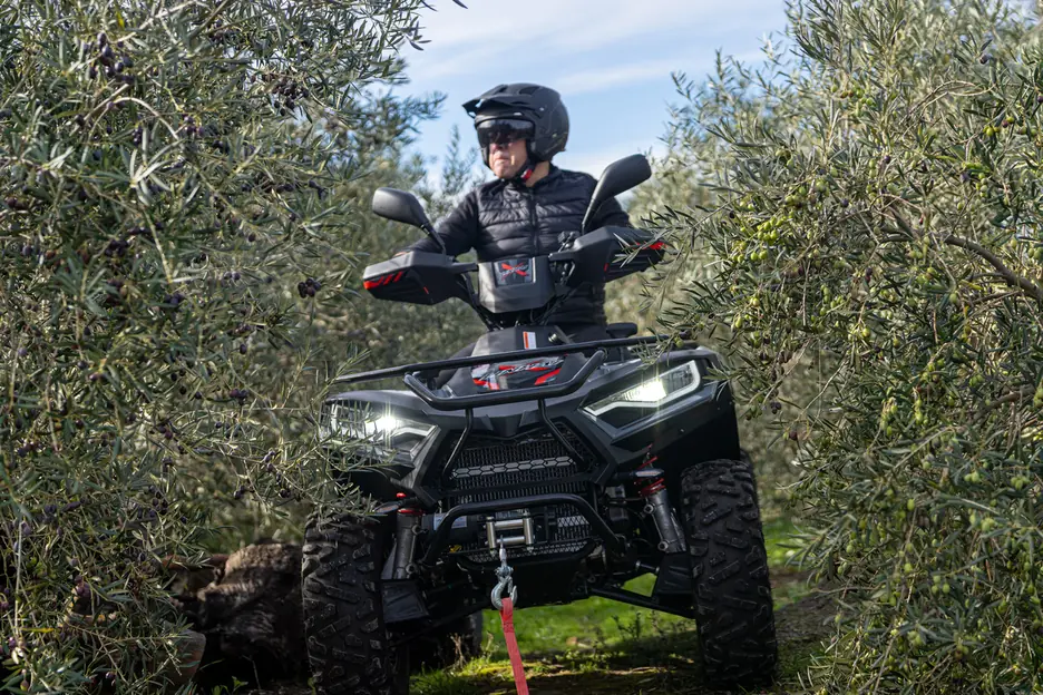 Conductor amb casc dalt d'un quad Linhai negre entre oliveres, amb el cable vermell del cabestrant enganxat al davant