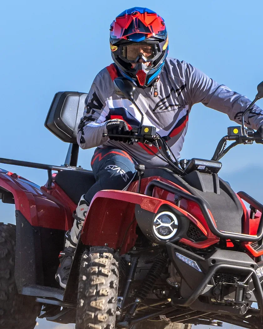 Conductor amb casc pilotant un quad Linhai vermell i negre, amb el cel blau i les muntanyes difuminades al fons