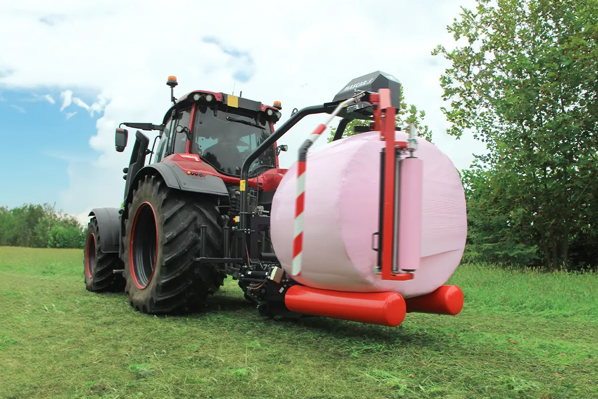 Encintadora Mascar vermella darrere d'un tractor vermell, amb una bala embolicada amb film rosa, en un prat