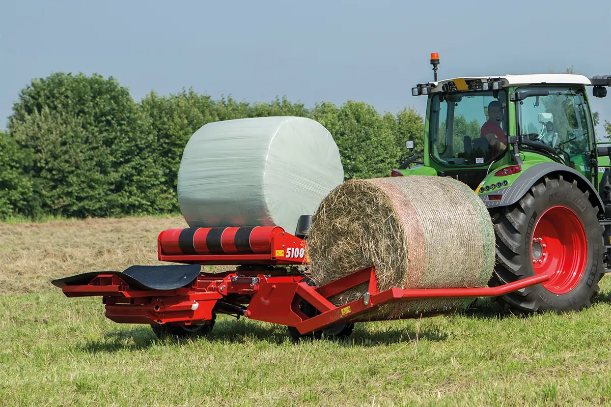 Encintadora Mascar 5100 remolcada darrere d'un tractor verd, amb una bala embolicada amb film blanc sobre els corrons i una altra d'herba a la forquilla lateral
