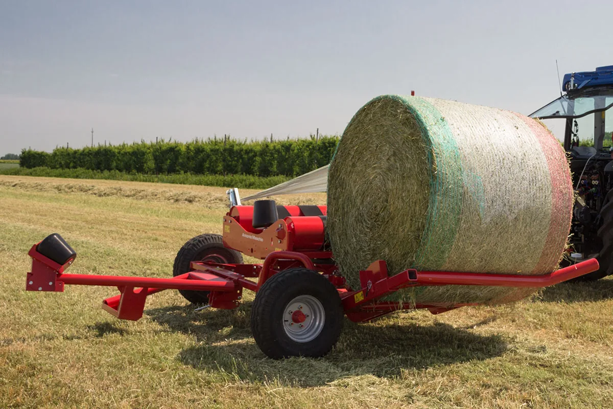 Encintadora Mascar vermella remolcada amb una bala d'herba sobre el braç de càrrega, abaixat al costat, i un tractor blau a la dreta, en un prat segat