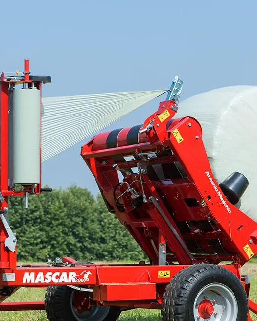 Encintadora Mascar vermella embolicant una bala rodona amb film blanc en un camp, amb la roda d'un tractor a l'esquerra
