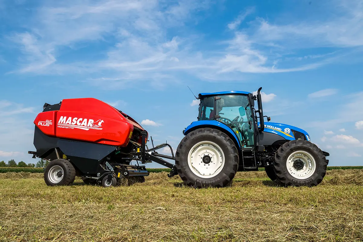 Rotoempacadora Mascar vermella enganxada a un tractor blau, de perfil, en un camp d'herba segada