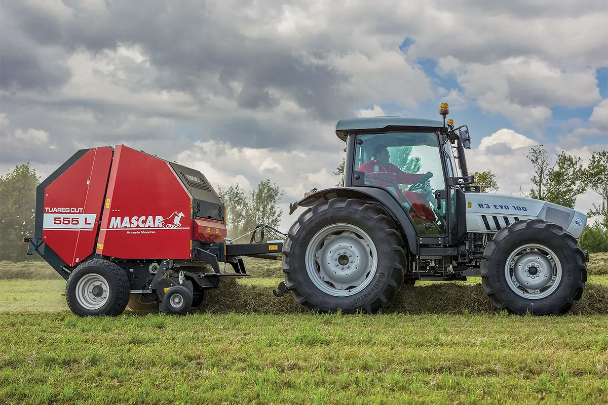 Rotoempacadora Mascar Tuareg Cut 555 de perfil darrere d'un tractor gris, sobre l'andana en un prat, sota un cel ennuvolat