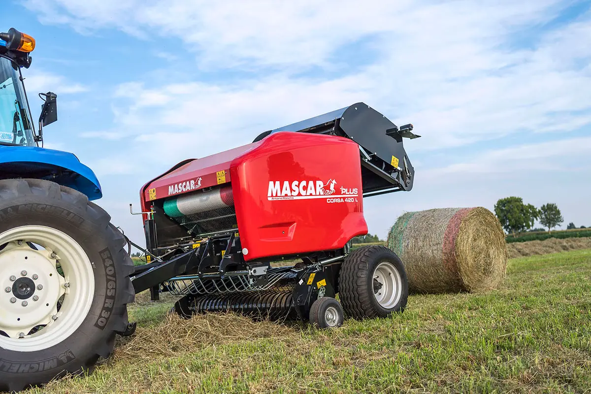 Rotoempacadora Mascar vermella darrere d'un tractor blau, amb una bala acabada de deixar en un prat segat