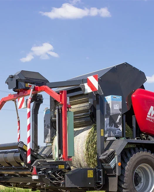 Rotoempacadora encintadora Mascar vermella treballant en un prat segat darrere d'un tractor verd, amb el cel blau i núvols al damunt