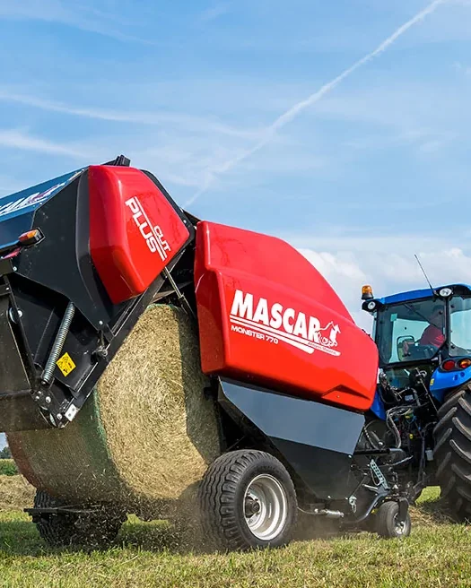 Rotoempacadora Mascar vermella amb la comporta oberta deixant anar una bala rodona en un camp segat, enganxada a un tractor blau, amb el cel blau al damunt