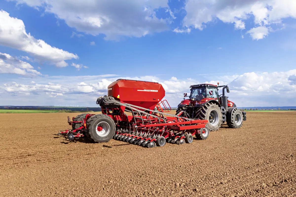Sembradora Horsch Avatar SD de tres quarts, enganxada a un tractor vermell sobre terra preparada, amb el cel blau i núvols al damunt