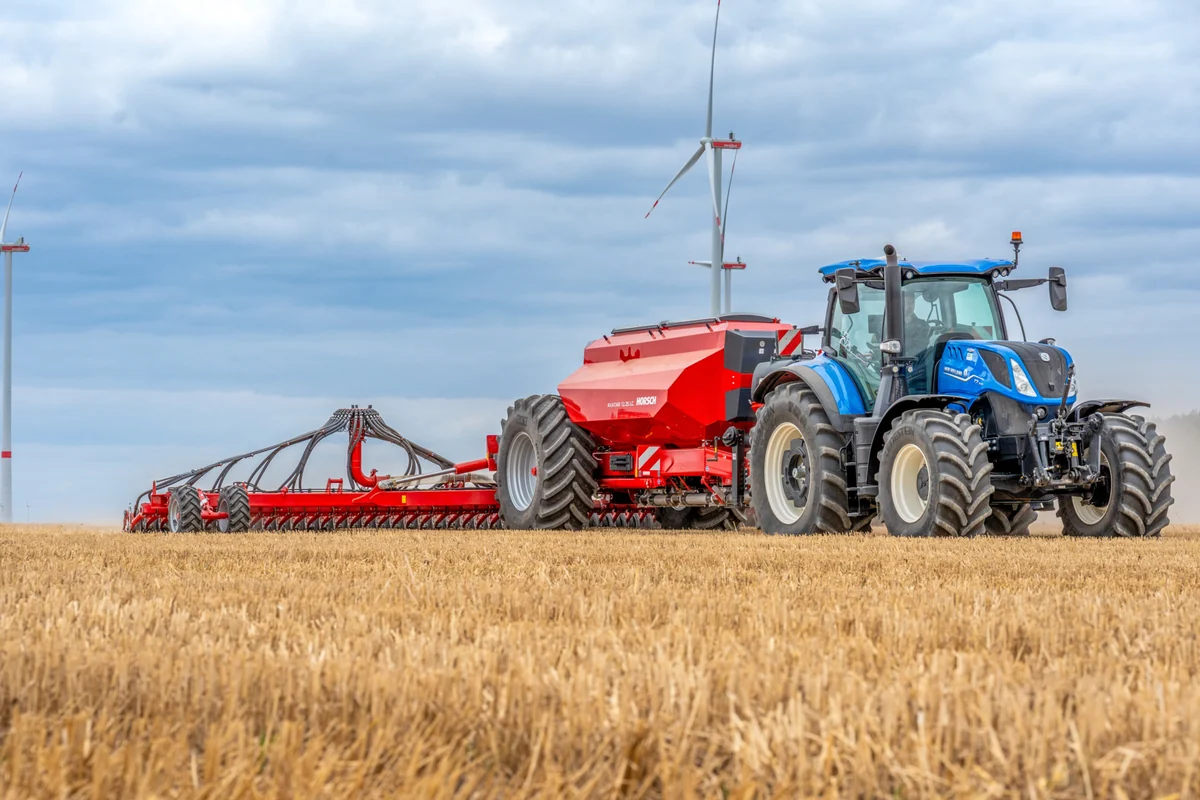 Sembradora Horsch Avatar LC darrere d'un tractor blau, en un camp de rostoll amb un aerogenerador al fons