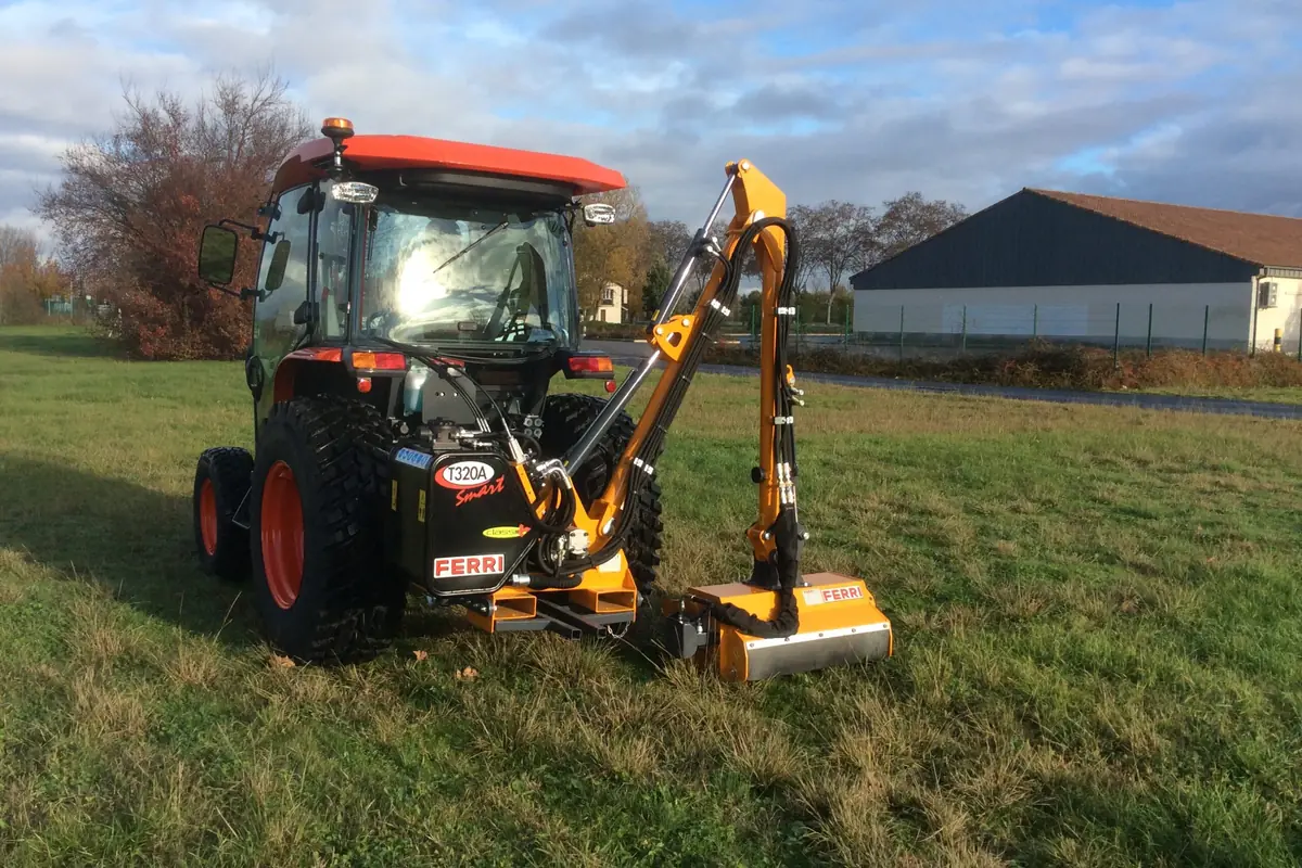 Tractor compacte amb un braç desbrossador Ferri groc segant l'herba d'un marge, amb una nau al fons