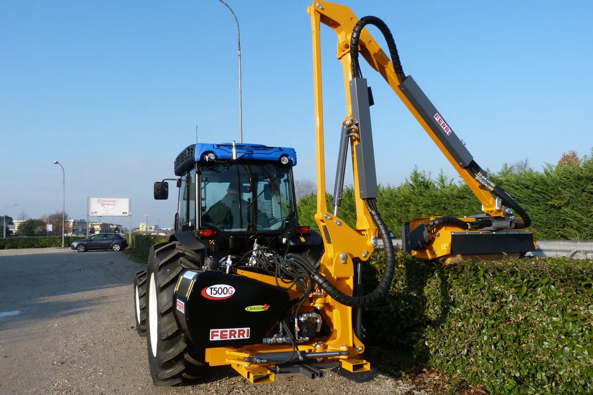 Tractor blau amb un braç desbrossador Ferri retallant una tanca vegetal a la vora d'una carretera