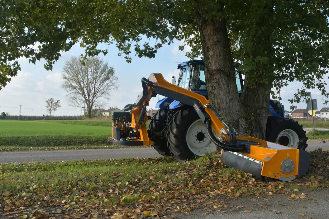 Tractor blau amb un braç desbrossador Ferri segant sota un arbre gran, al costat d'una carretera