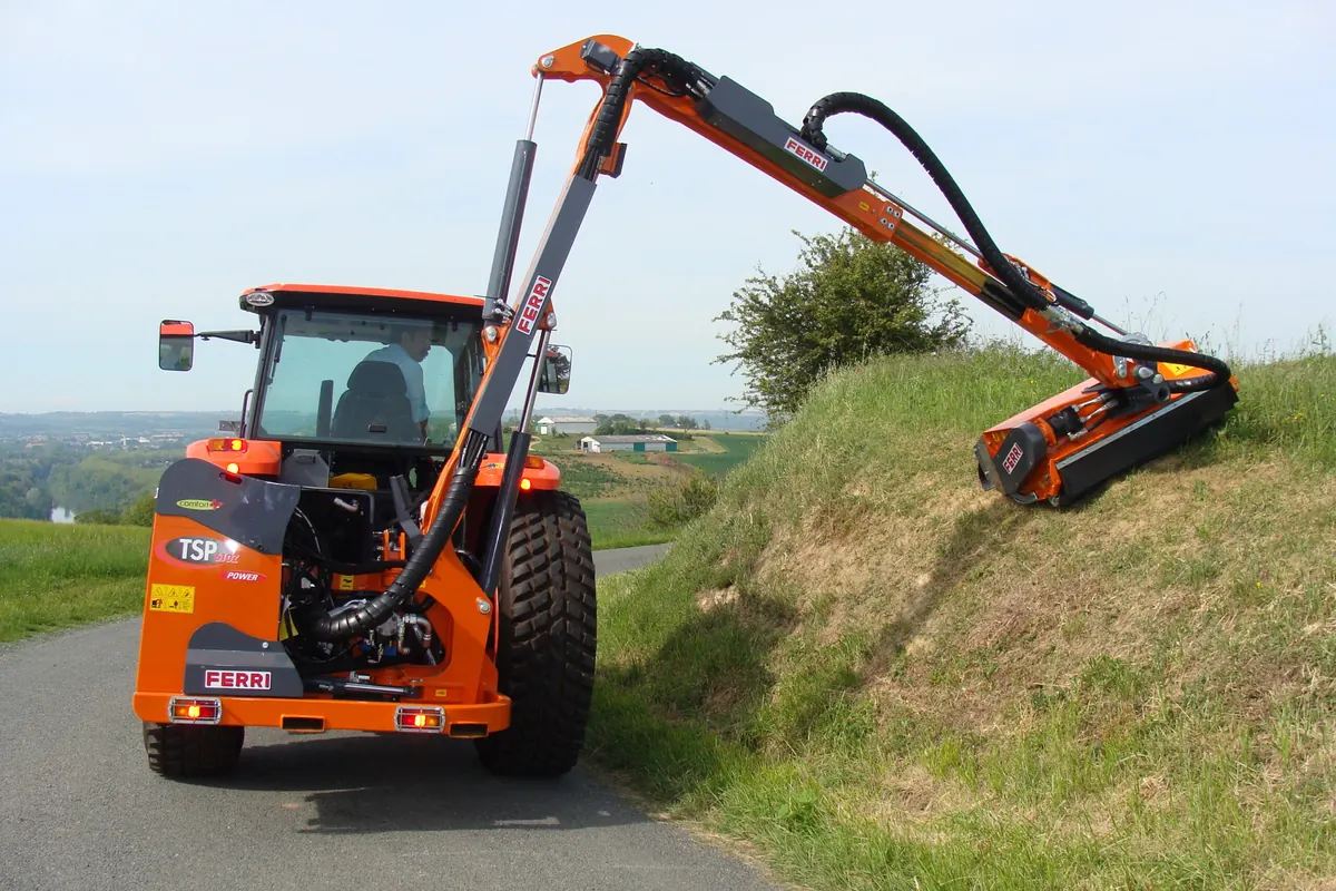 Braç desbrossador Ferri TSP 610 Z muntat al darrere d'un tractor taronja, segant el talús d'una carretera estreta, vist des del darrere